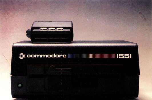 Eine schwarze Floppy-Diskettenstation mit der Aufschrift »commodore 1551« und farbigem Regenbogen-Logo, dunkler Hintergrund