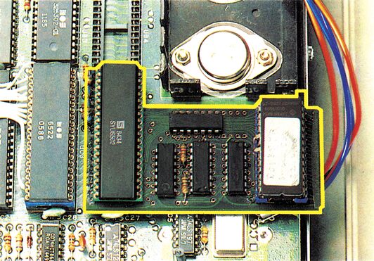 Nahaufnahme einer bestückten Platine mit ICs und Chips; ein Bereich ist gelb umrandet, bunte Kabel führen nach rechts
