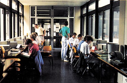 Innenraum mit mehreren Personen an Tischen mit Computern und Monitoren entlang einer Fensterfront