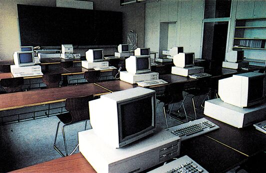 Computerraum mit einer Reihe von Personal-Computern mit Monitoren und Tastaturen auf langen Tischen, im Hintergrund eine große dunkle Tafel und Regale