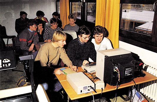 Gruppe junger Personen, die dicht gedrängt um einen Tisch mit Computer, Monitor und Diskettenlaufwerk stehen und sitzen