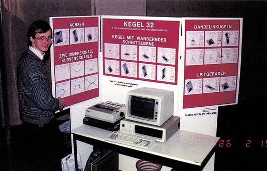 Ein Mann steht an einem Ausstellungsstand mit Stellwand und Postern (»ACHSEN«, »KEGEL 32«, »DANDELINKUGELN«), davor ein Computer mit Monitor und Tastatur
