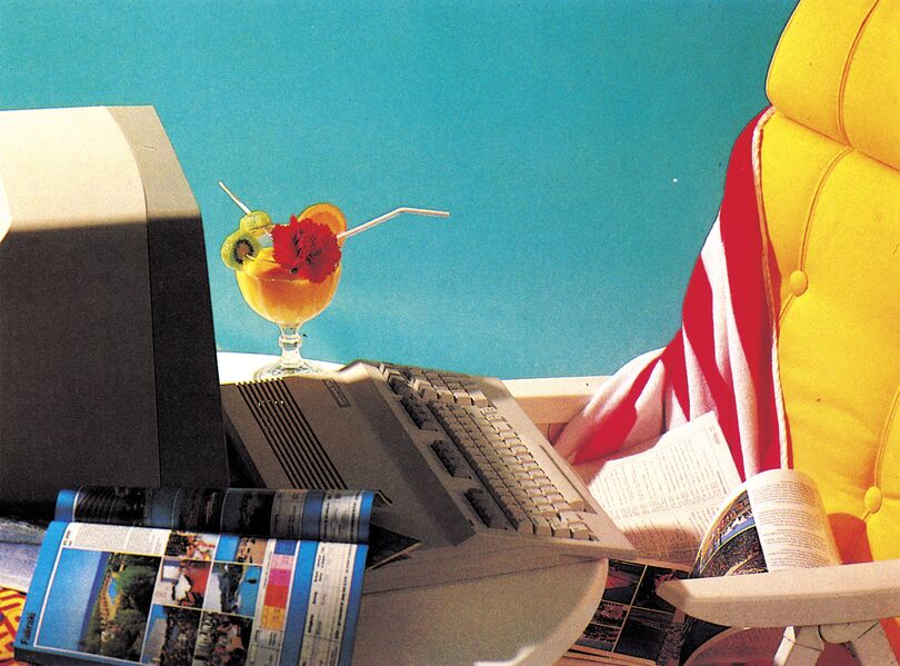 Ein Computer mit Tastatur und Monitor im Freien, darauf ein Cocktailglas mit Früchten, daneben ein gelber Liegestuhl mit rot-weiß gestreiftem Tuch