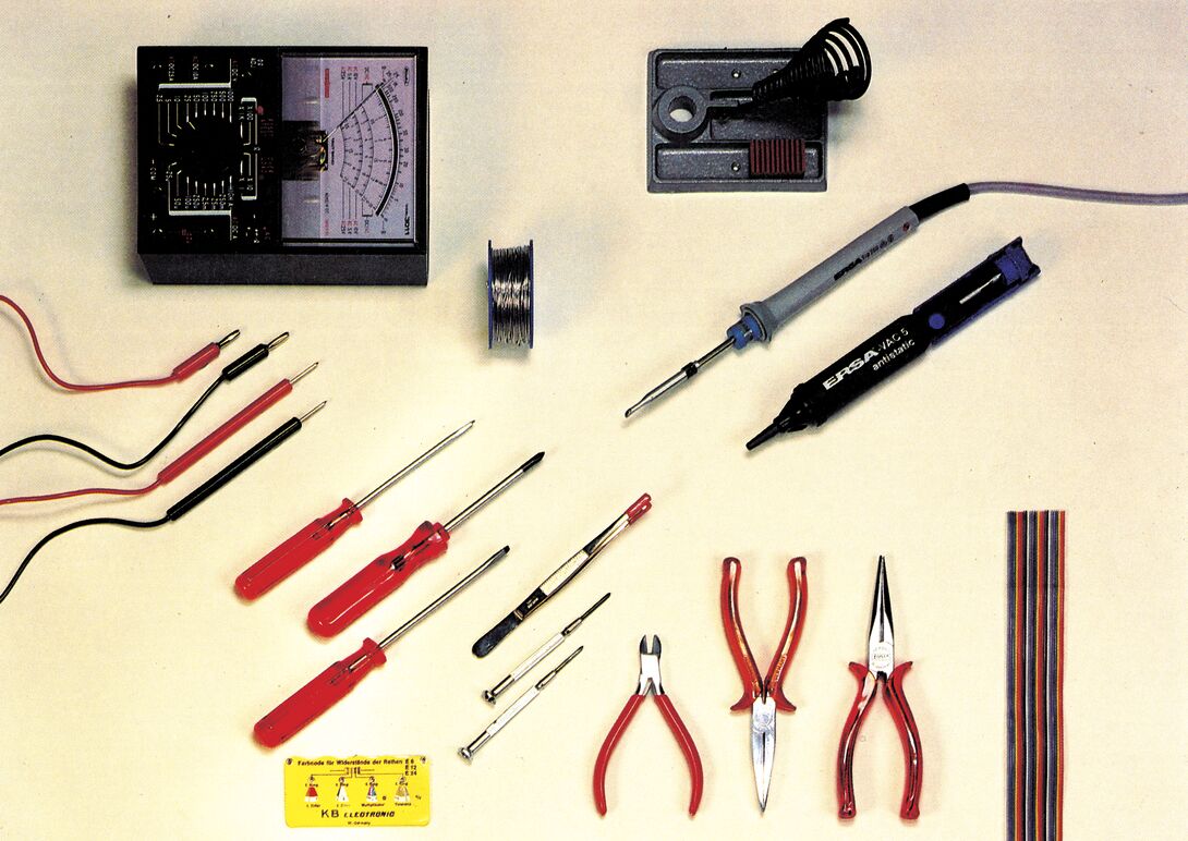 Aufsicht auf Werkzeuge und Meßgeräte auf hellem Untergrund: ein analoges Multimeter mit Meßleitungen, Lötkolben mit Ablage, Entlöt-Saugpumpe, Lötzinn, Schraubendreher, Pinzette, Zangen, Seitenschneider, ein Kärtchen mit Farbcode-Tabelle und ein Flachbandkabel