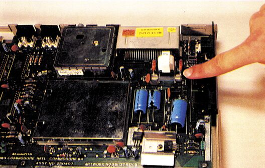 Geöffnetes C-64-Gehäuse mit Blick auf die Hauptplatine mit Chips und Bauteilen; ein Finger zeigt von rechts auf eine Stelle der Platine