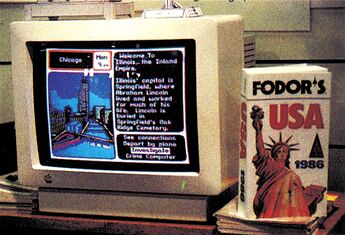 Foto eines Monitors mit einem Spielbildschirm neben einem Reiseführer-Buch »FODOR'S USA 1986« mit Freiheitsstatue
