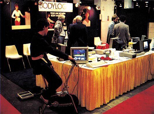 Messestandfoto: eine Person auf einem Heimtrainer/Ergometer vor einem Stand mit Schriftzug »BODYLOG«, im Hintergrund Menschen und Computer