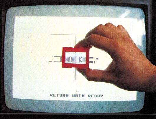 Hand hält eine kleine rote Plastiklinse vor einen Fernsehbildschirm, auf dem »OK« steht