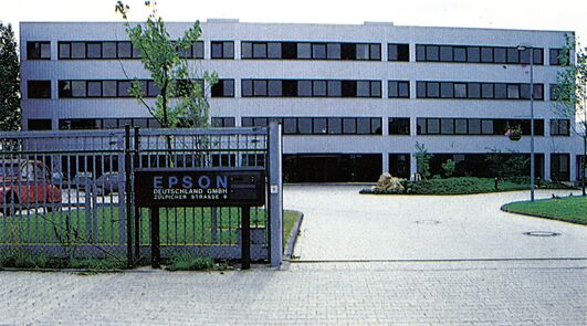 Farbfoto eines mehrstöckigen Bürogebäudes hinter einem Zaun mit dem Firmenschild »EPSON Deutschland GmbH«