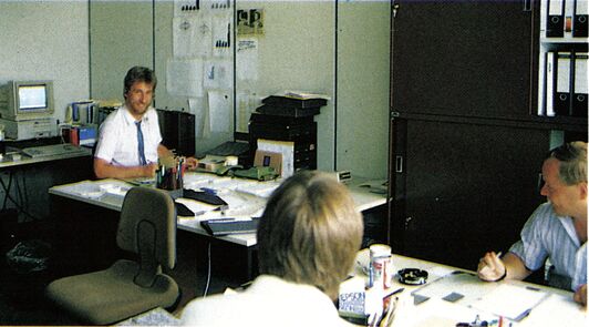 Farbfoto eines Büroraums, in dem mehrere Personen an einem Tisch mit Computer und Telefon sitzen