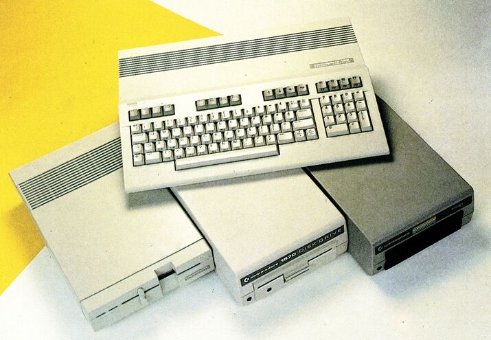 Drei Diskettenlaufwerke nebeneinander, dahinter eine C-128-Tastatureinheit, auf gelb-weißem Hintergrund