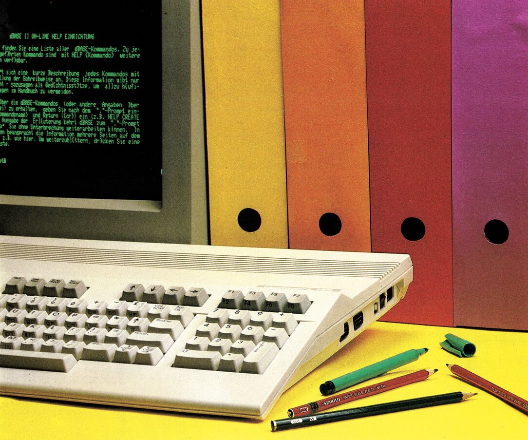 Farbfoto einer C-128-Tastatur und eines Monitors vor bunten Ordnerrücken; auf dem grünen Bildschirm der dBASE-II-Hilfetext