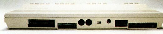 Rückseite eines flachen Computergehäuses mit langem Steckplatz, einer Buchsenleiste, zwei runden Buchsen, einem kleinen Schalter und zwei rechteckigen Öffnungen